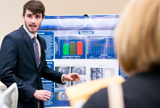 a student in a suit points to a poster with graphs