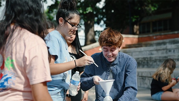 A group of students outdoors conducts a science experiment using a funnel and container, with one student smiling while pouring material into the setup as others observe.