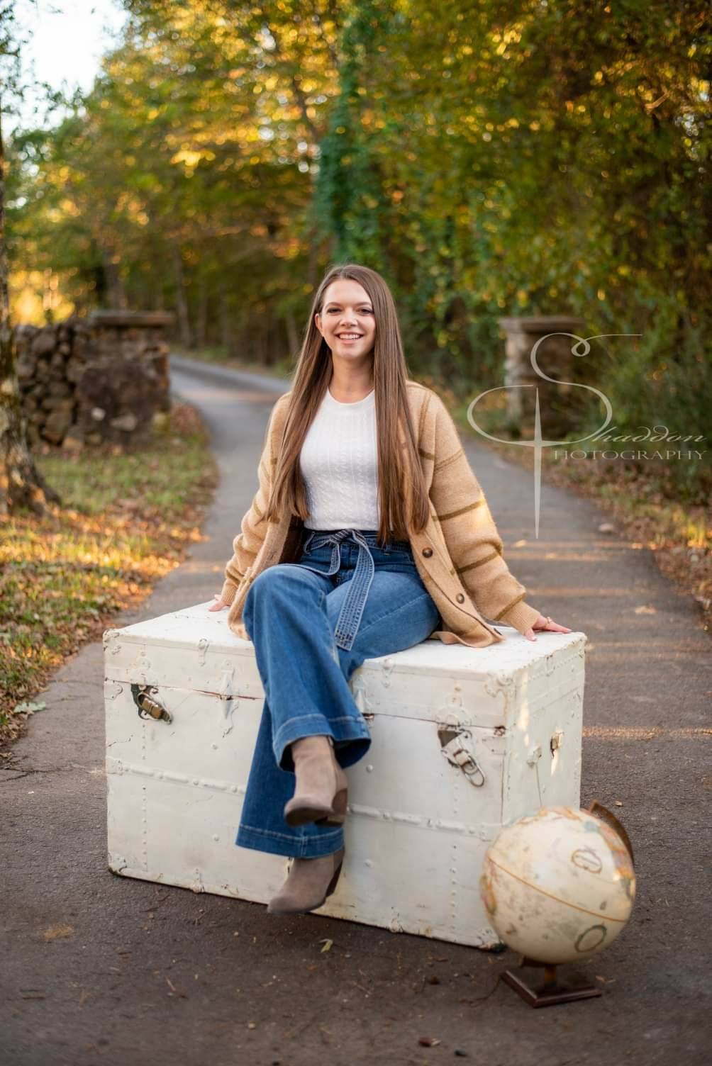 Avery Powell, smiling while sitting on a white, antique trunk. A globe sits on the ground by the trunk.