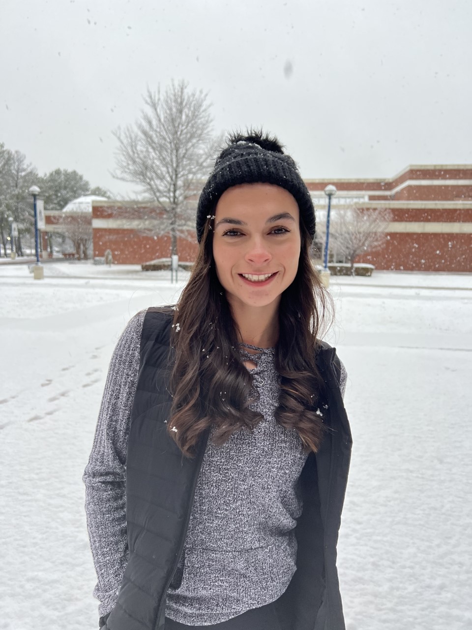 The UAFS campus is covered in snow. Isabella stands in winter clothing near the campus center. She is smiling. 