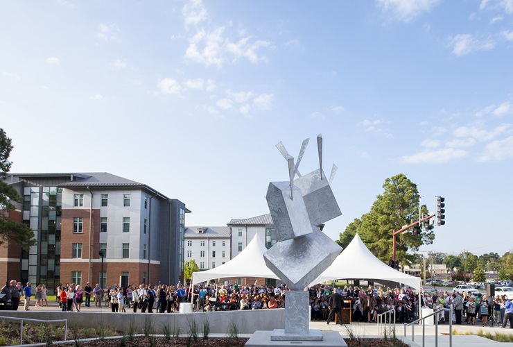 Crowd gathered on the UAFS campus around a public art sculpture during an outdoor event, with campus buildings and tents in the background.