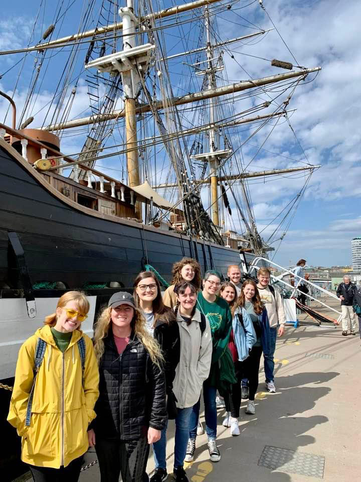 Ten people stand in a line in front of a historic ship. 
