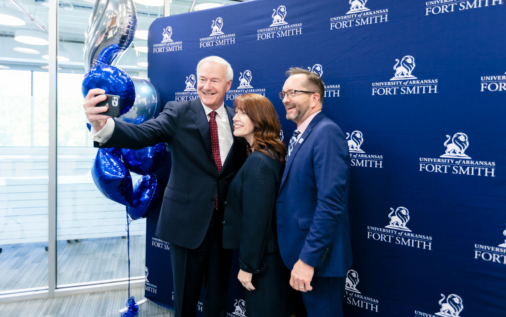 Governor Asa Hutchinson, UAFS Chancellor Terisa Riley, and CED Executive Director, Kendall Ross pose for a selfie in front of a UAFS backdrop. 