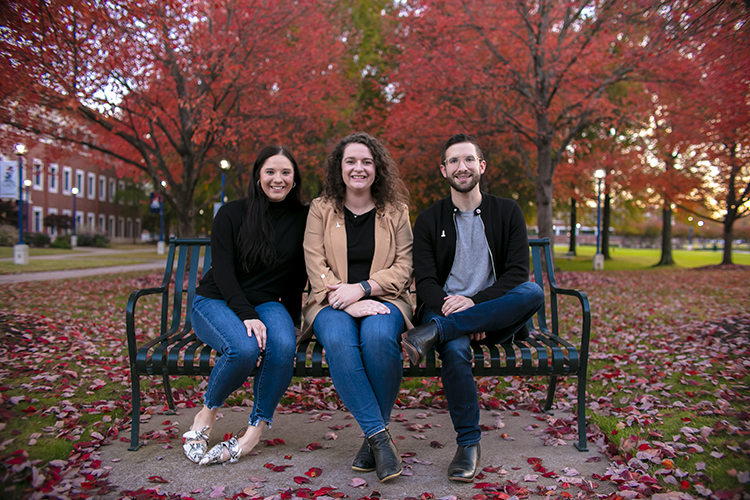 Three university staff members sit on a campus bench surrounded by trees during the fall.