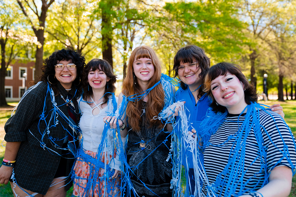 A group of students smile on the UAFS Campus Green covered in blue and white streamer confetti.