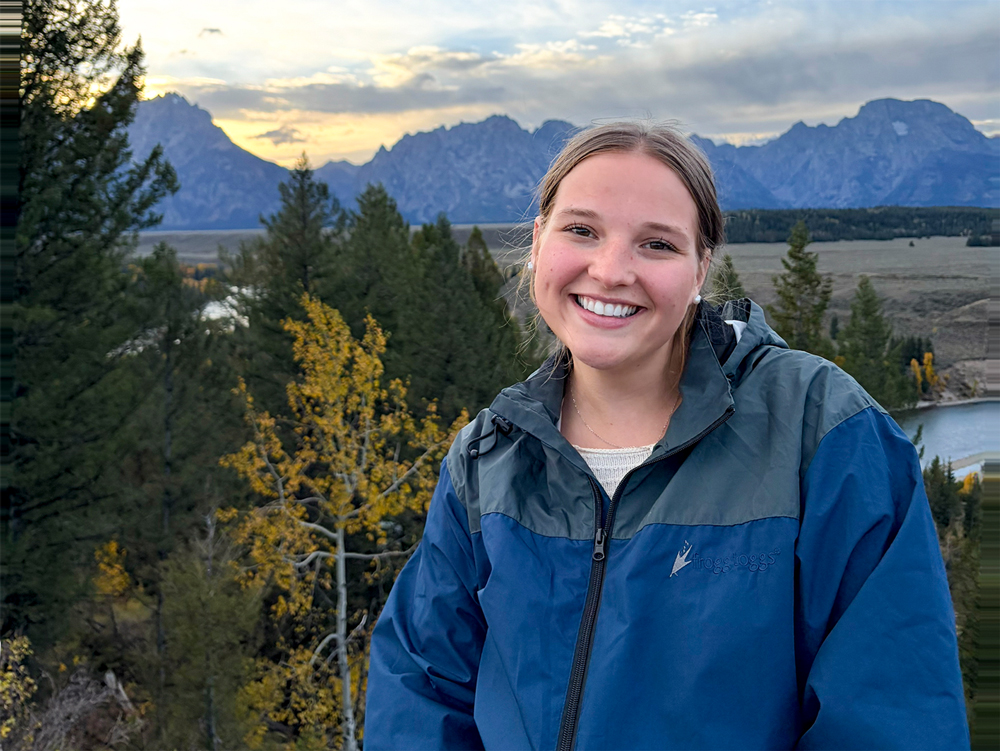 Ella Driscoll snaps a picture out west with pine trees and mountains with a setting sun behind her.