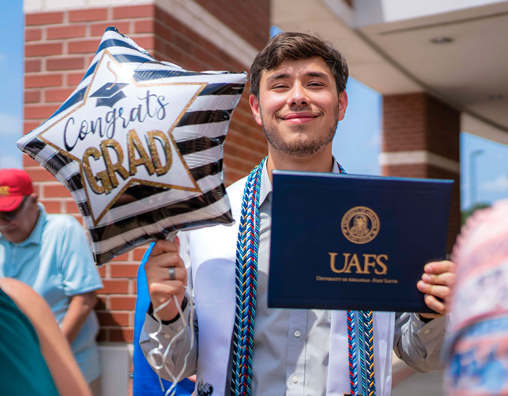 UAFS graduate smiles for the camera holding his diploma and a "Congrats Grad" star-shaped balloon