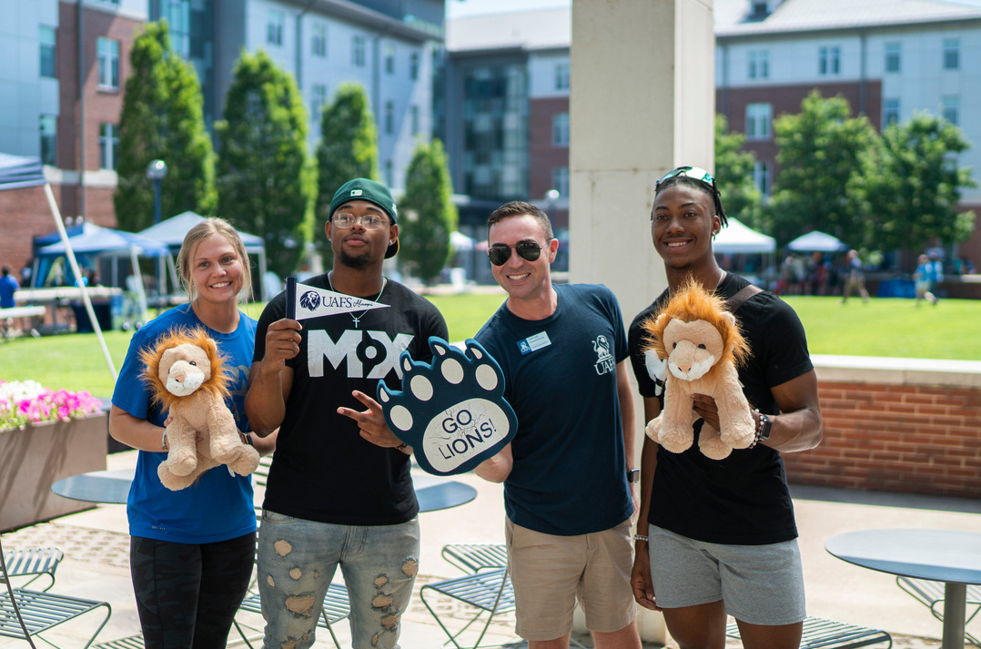 four students hold stuffed lions and swag in front of the UAFS dorms