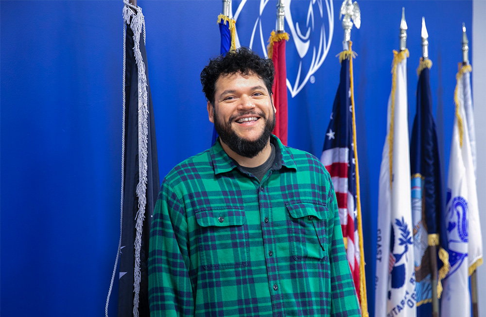 Raul Tabares, a senior business administration student at UAFS, stands in front of flags denoting the U.S. military branches at the UAFS Veterans Resource Center