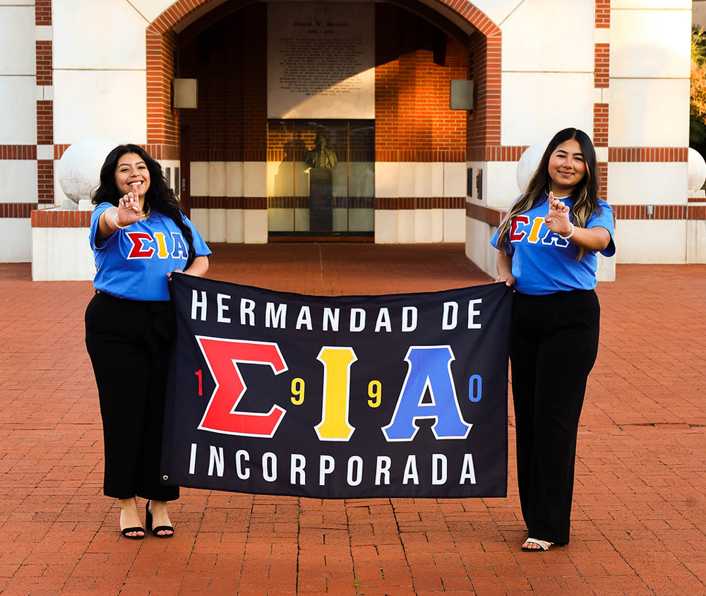 Jaylin Barroso (left) and Kimberly Villagrana stand in front of the UAFS Bell Tower holding a flag for Hermandad de Sigma Iota Alpha, Inc., or SIA, the newest sorority to UAFS.