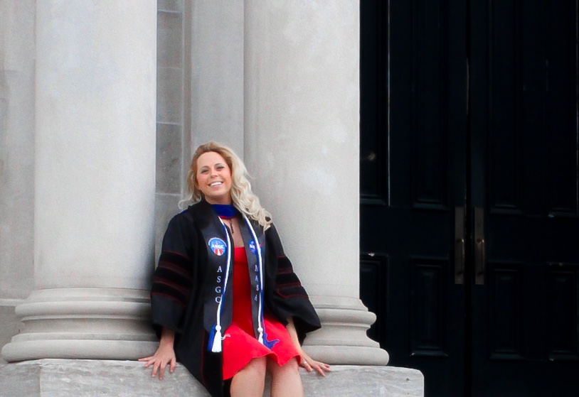 Woman in graduation robes sitting besides pillars leading into a university building